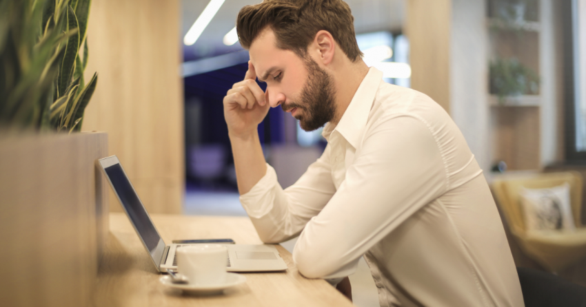 Man holds hand to head as he frowns at a laptop with a cup of coffee nearby